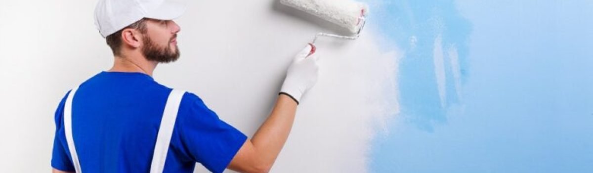 Back view of handsome young painter in white dungarees, blue t-shirt, cap and gloves painting a wall with paint roller.